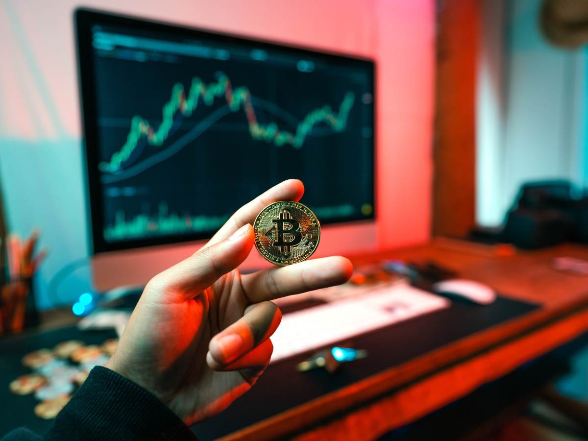  Bitcoin u ruci ispred monitora sa grafikonom cena 